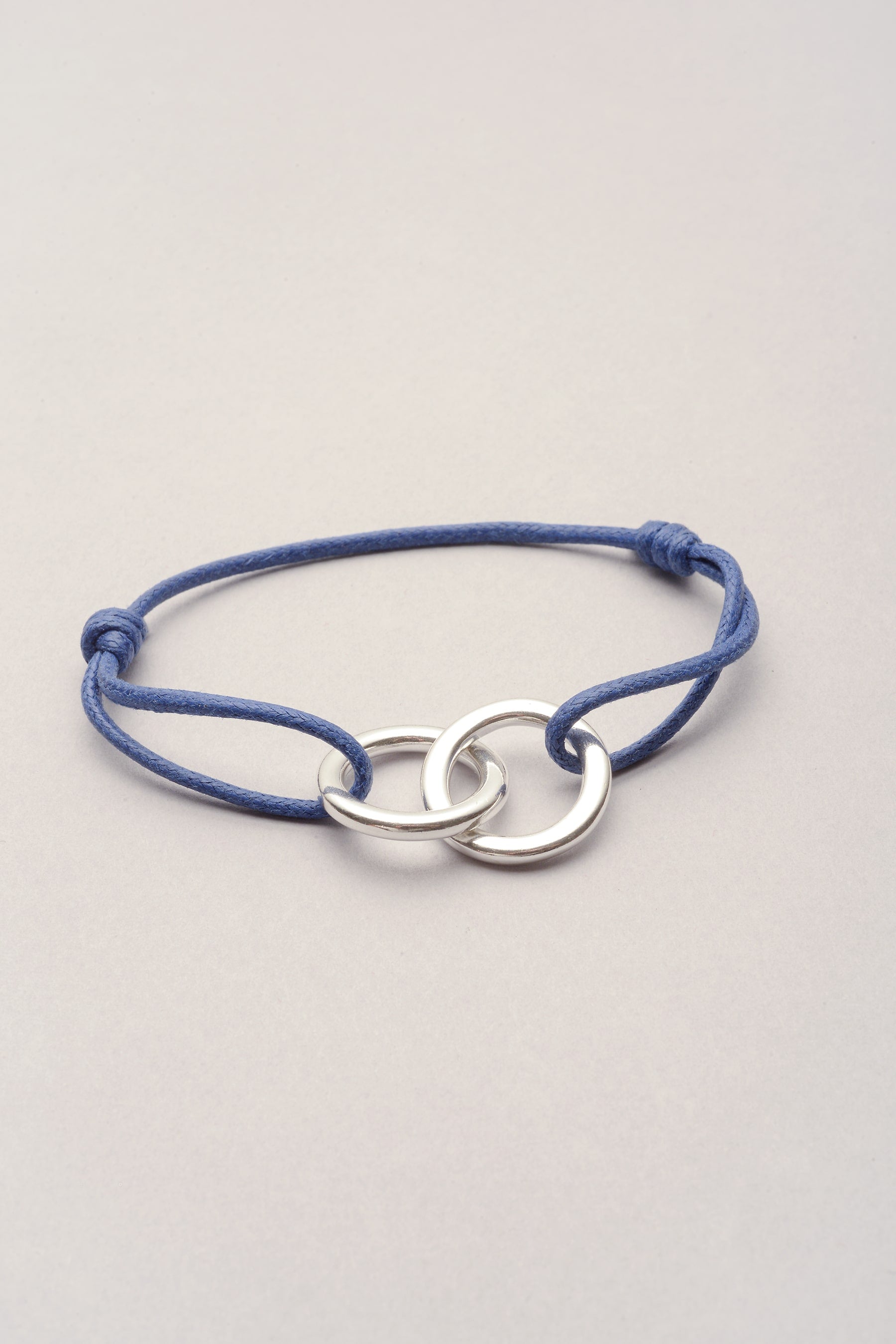 Le bracelet est composé d'un pendentif représentant deux anneaux entrelacés. Réglable, il est possible d'ajuster le cordon de coton coloré grâce à ses deux noeuds coulissants. 
