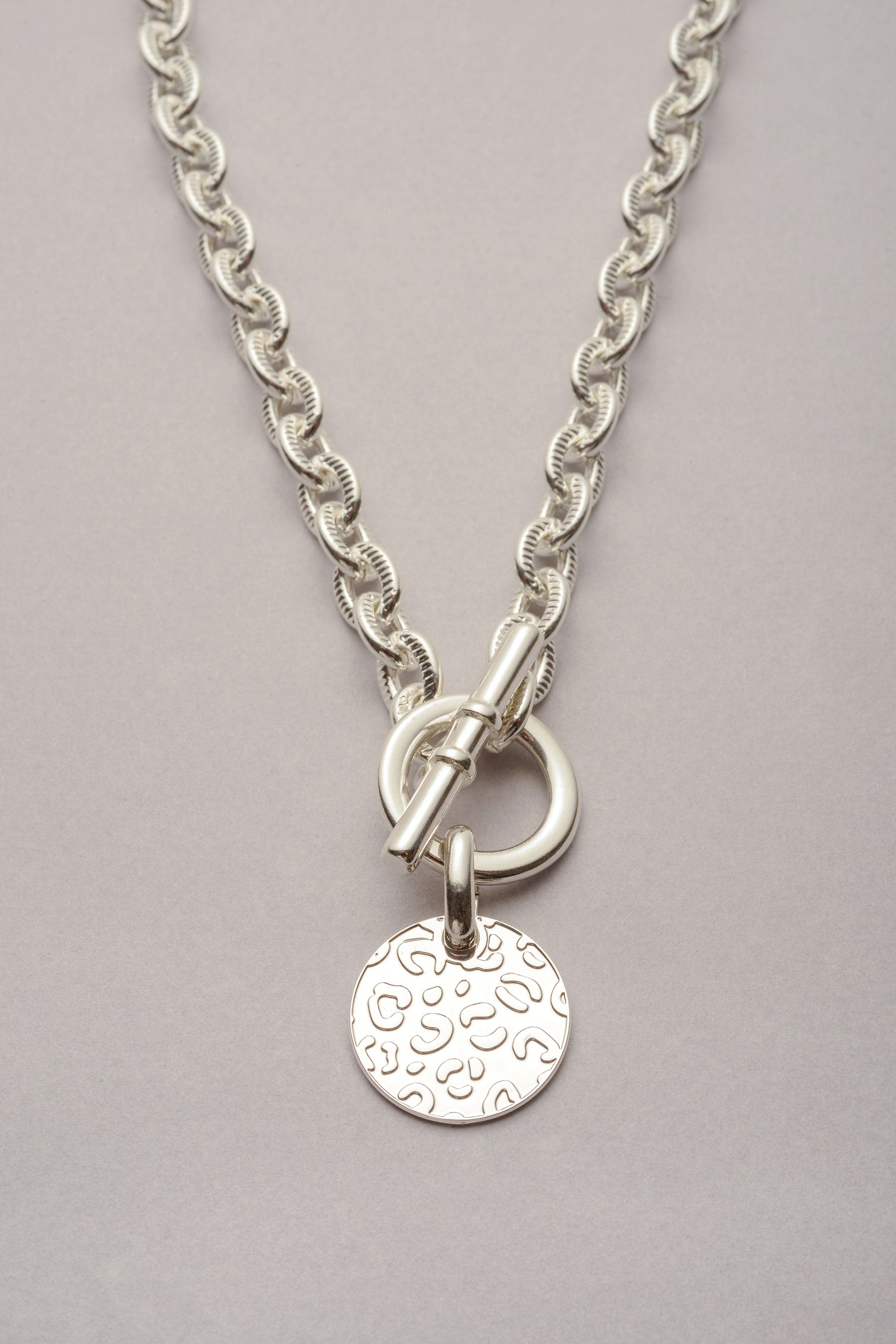 Collier Scarlett Médaille Léopard argenté. Ce bijou est composé d'une chaîne striée et d'une médaille avec des reliefs léopard. Pratique, il s'ouvre et se ferme grâce à un fermoir en T. 